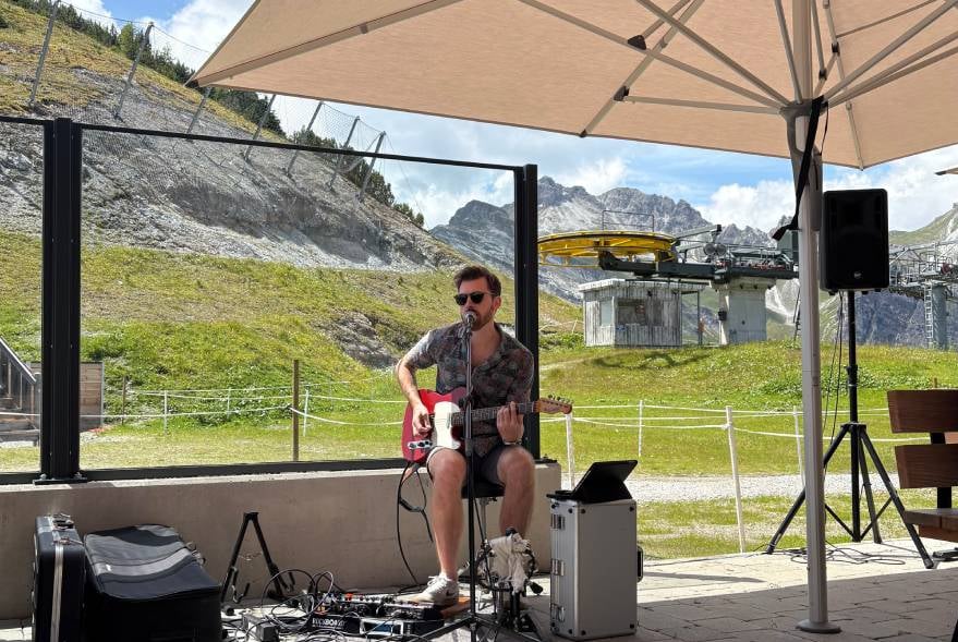 Person sitzt, spielt Gitarre und singt auf der Terrasse des Panorama Restaurant Kreuzjoch unter einem Sonnenschirm